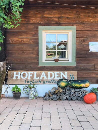 A rustic farm shop with a wooden sign and a cozy atmosphere. In front of the shop, there are wooden logs and decorative pumpkins. | © Kurkommission Bad Blumau