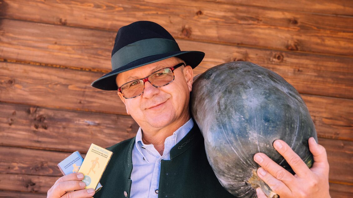 Ein Mann in traditioneller Kleidung hält einen großen Kürbis und Tickets in der Hand. Im Hintergrund sieht man eine Holzwand. | © Kurkommission Bad Blumau