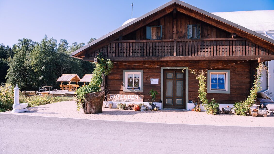 Ein traditionelles Holzhaus mit einem Balkon und bunten Blumen vor der Tür. Im Hintergrund sind Bäume und eine klare blaue Himmel sichtbar. | © Kurkommission Bad Blumau