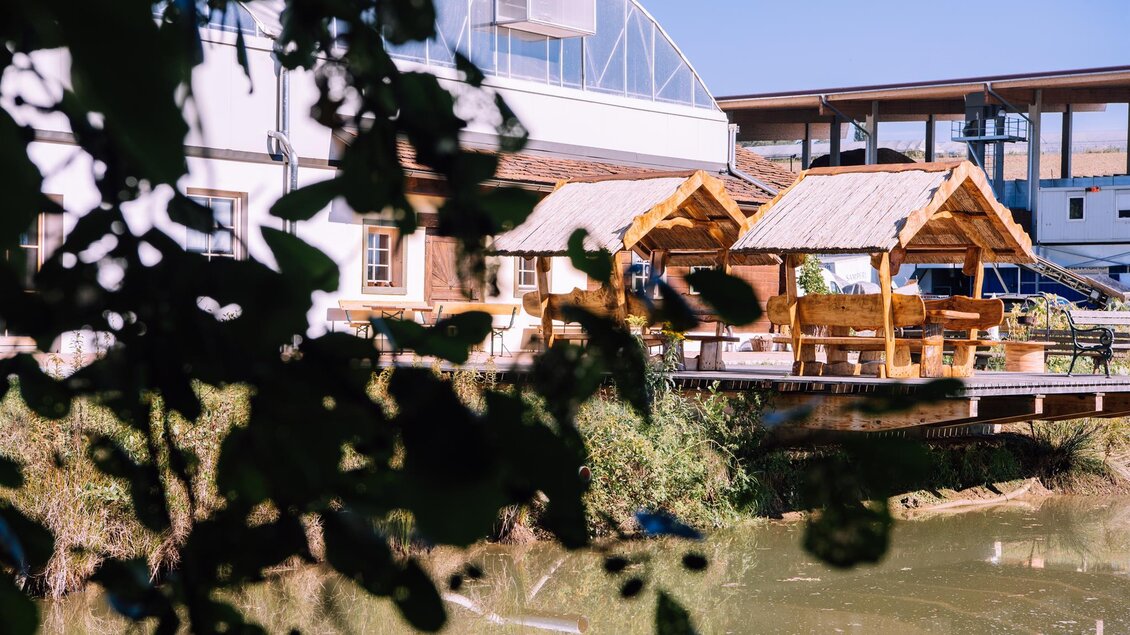 Ein ruhiger Ausblick auf ein Gebäude mit einem kleinen Teich. Im Vordergrund sind überdachte Sitzgelegenheiten zu sehen. | © Kurkommission Bad Blumau