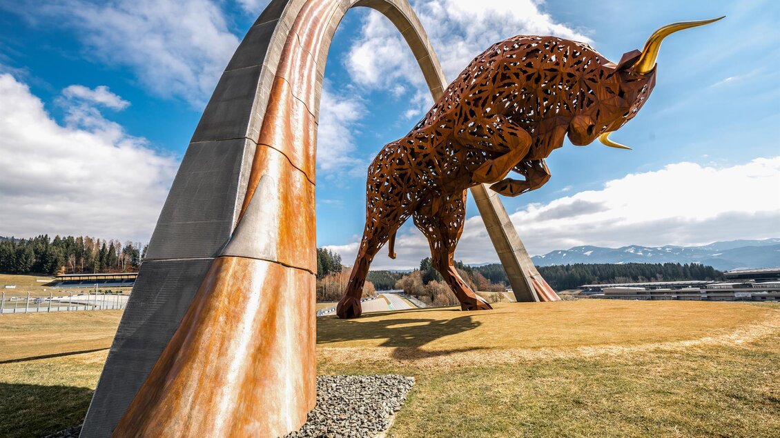 Eine große, kunstvolle Skulptur eines Bullen steht unter einem bogenförmigen Monument. Die Landschaft ist grün mit einer klaren blauen Himmel im Hintergrund. | © Andreas Berr - Region Spielberg