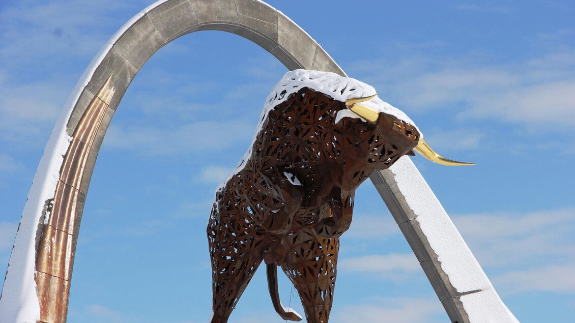 Eine beeindruckende Skulptur eines Bullen steht unter einem großen Bogen. Der Bulle hat große Hörner und ist teilweise mit Schnee bedeckt. | © Anita Fössl