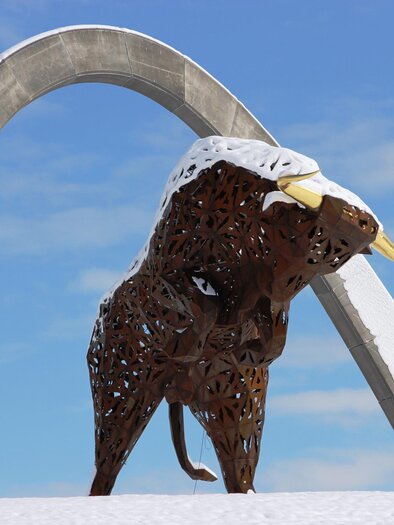 An impressive sculpture of a bull stands under a large arch. The bull has large horns and is partially covered in snow. | © Anita Fössl