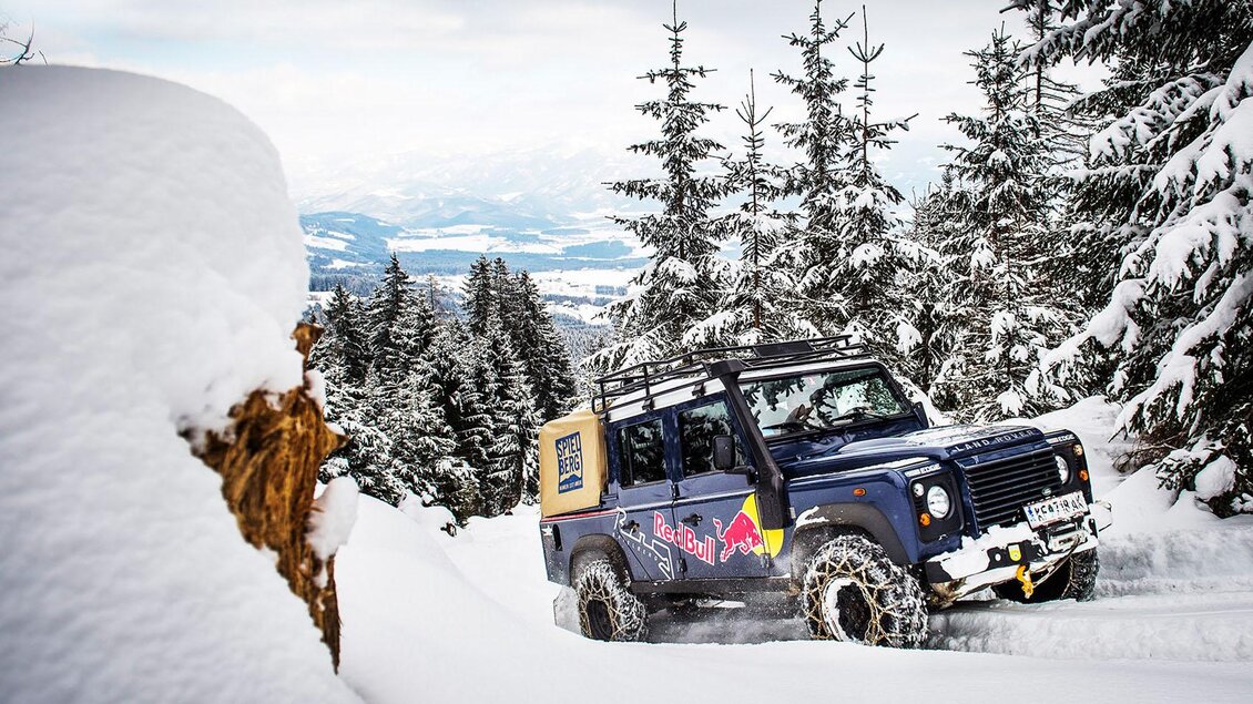 Ein Geländewagen fährt durch eine verschneite Landschaft mit hohen Tannenbäumen. Im Hintergrund sind sanfte Hügel und ein klarer Himmel zu sehen. | © Lucas Pripfl