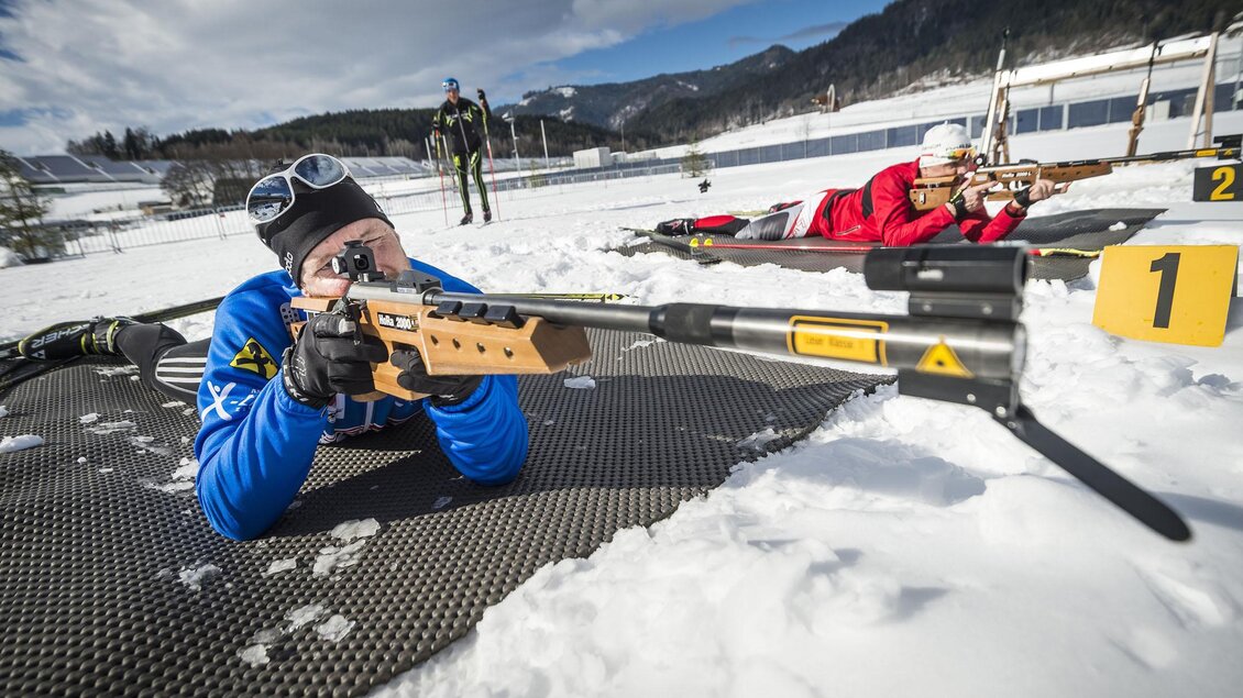 Ein Skisportler liegt im Schnee und zielt mit einem Gewehr. Im Hintergrund sind weitere Schützen zu sehen, umgeben von einer winterlichen Landschaft. | © Philip Platzer