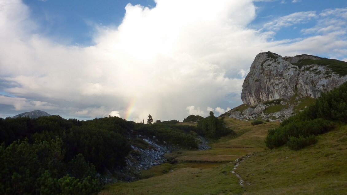 Eine malerische Landschaft mit grünen Wiesen und einem großen Felsen. Im Hintergrund sind Wolken und ein leichter Regenbogen zu sehen.