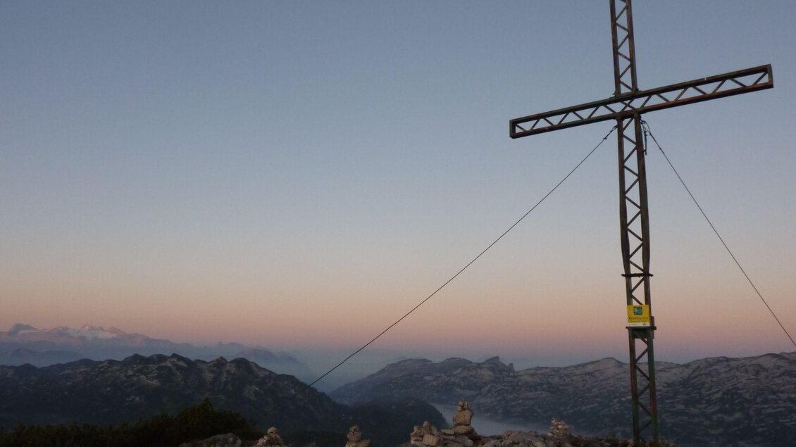 Ein großes Kreuz auf einem Berggipfel mit einer sanften Hügelkette im Hintergrund. Der Himmel zeigt sanfte Farben während des Sonnenuntergangs.