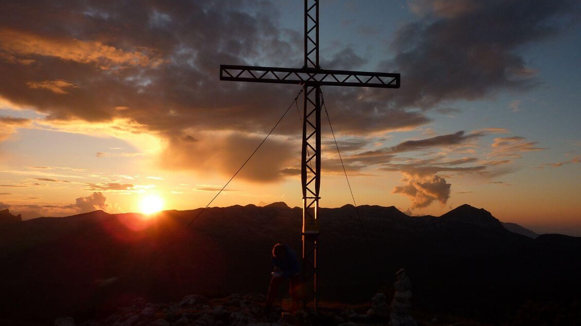 Ein hochwertiges Bergpanorama bei Sonnenuntergang mit einem großen Kreuz im Vordergrund. Die Berge sind im Hintergrund sichtbar, und der Himmel hat schöne orange und blaue Farbtöne.