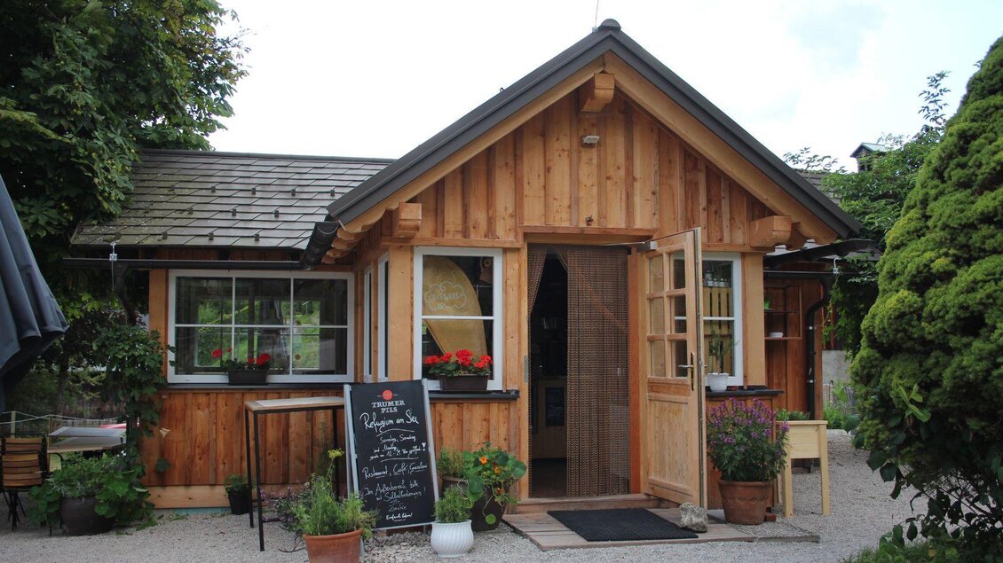 Ein charmantes Holzhaus mit einem einladenden Eingang. Vor dem Haus stehen Pflanzen und eine Tafel mit Angeboten. | © Viola Lechner