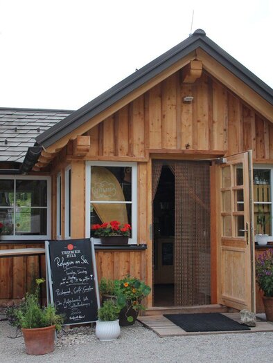 A charming wooden house with an inviting entrance. In front of the house, there are plants and a board with offers. | © Viola Lechner