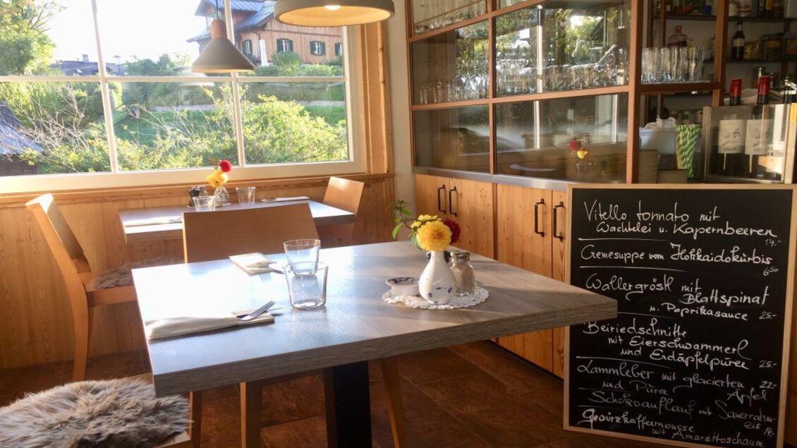 Ein gemütliches Restaurant mit Holzmöbeln und einem großen Fenster. Auf dem Tisch steht eine kleine Blumenvase und nebenan eine Tafel mit Speiseangeboten. | © Robert Hocker