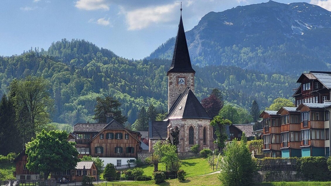 Ein malerisches Dorf am See mit einer Kirche und traditionellen Häusern. Im Hintergrund erheben sich grüne Berge unter einem blauen Himmel. | © Petra Kirchschlager