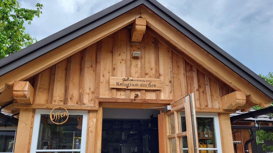 Ein rustikales Holzgebäude mit einer offenen Tür. Das Schild oben trägt die Aufschrift "Refugium am See". | © Petra Kirchschlager