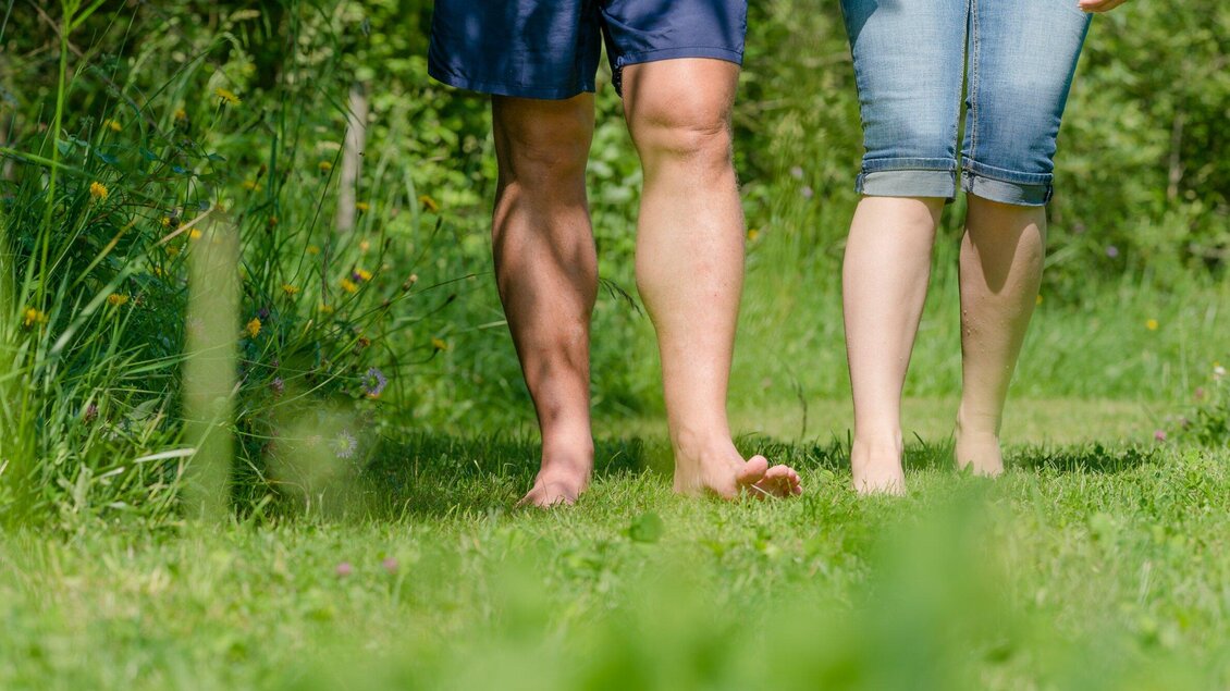 Zwei Personen gehen barfuß über eine grüne Wiese. Die Sonne scheint und die Umgebung ist naturbelassen. | © TV Region Graz - Mias Photoart