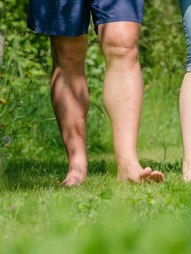 Zwei Personen gehen barfuß über eine grüne Wiese. Die Sonne scheint und die Umgebung ist naturbelassen. | © TV Region Graz - Mias Photoart