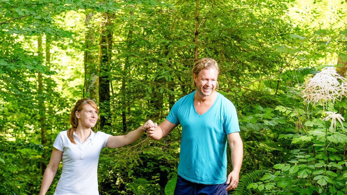 Ein Paar geht Hand in Hand durch einen grünen Wald. Die Sonnenstrahlen scheinen durch die Bäume und schaffen eine fröhliche Atmosphäre. | © TV Region Graz - Mias Photoart