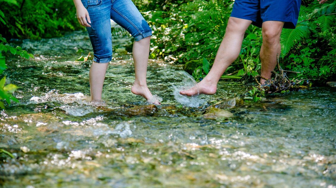 Zwei Personen stehen im Wasser eines kleinen Baches. Die Füße sind barfuß und es spritzen kleine Wassertropfen. | © TV Region Graz - Mias Photoart