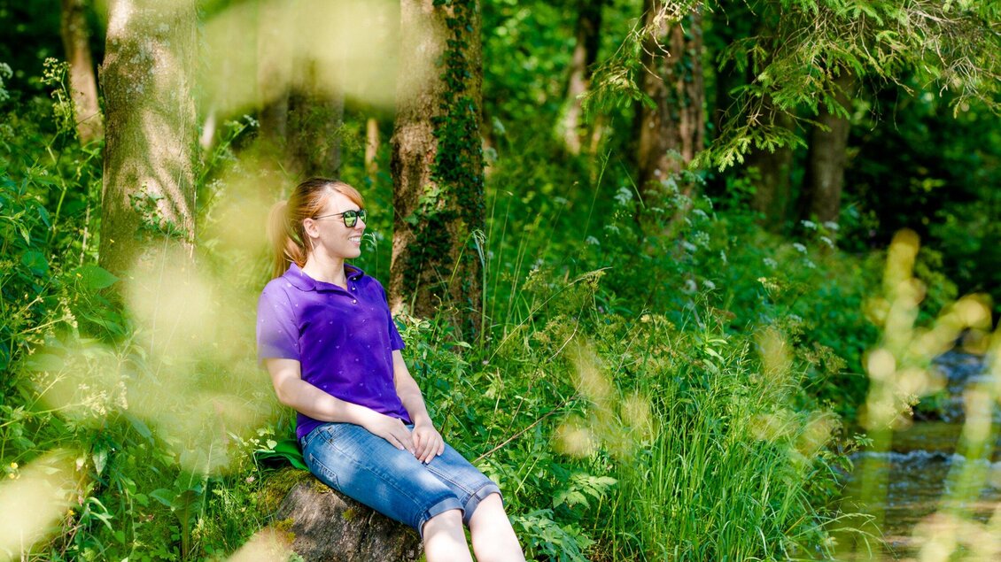 Eine Frau sitzt entspannt auf einem Baumstamm am Ufer eines Baches. Umgeben von grünen Pflanzen und Bäumen genießt sie die Natur. | © TV Region Graz-  Mias Photoart
