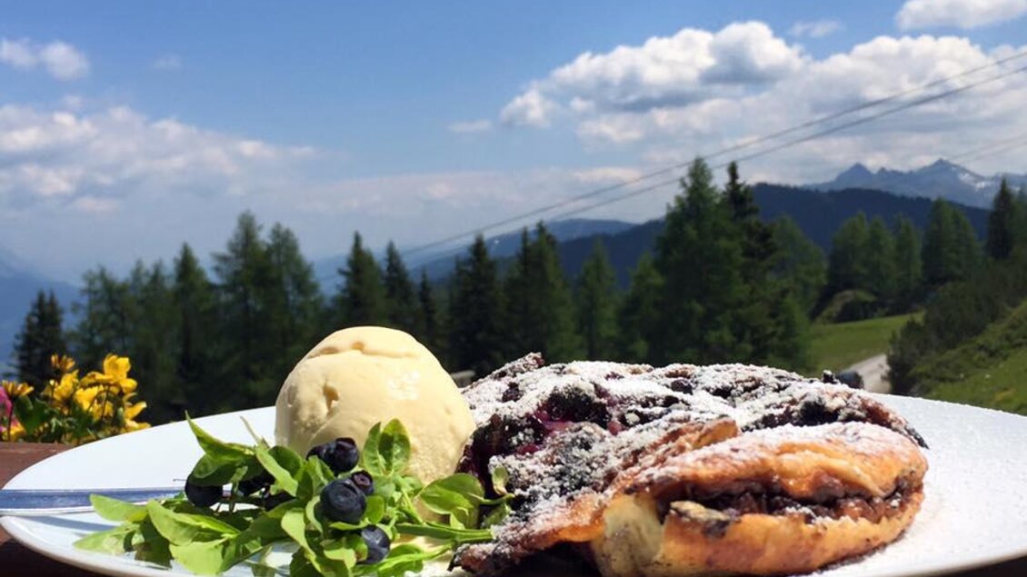 Ein köstliches Dessert mit einem Stück Gebäck, einer Kugel Eis und frischen Beeren. Im Hintergrund sieht man eine schöne Berglandschaft und einen blauen Himmel. | © Reiteralmhütte 