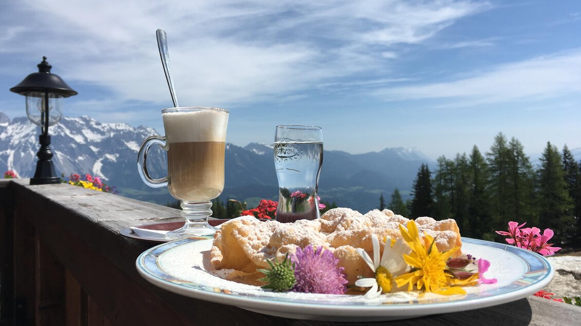 Ein Teller mit köstlichen Süßspeisen und bunten Blüten steht auf einem Tisch. Im Hintergrund erstreckt sich eine majestätische Berglandschaft unter einem blauen Himmel. | © Markus Trinker