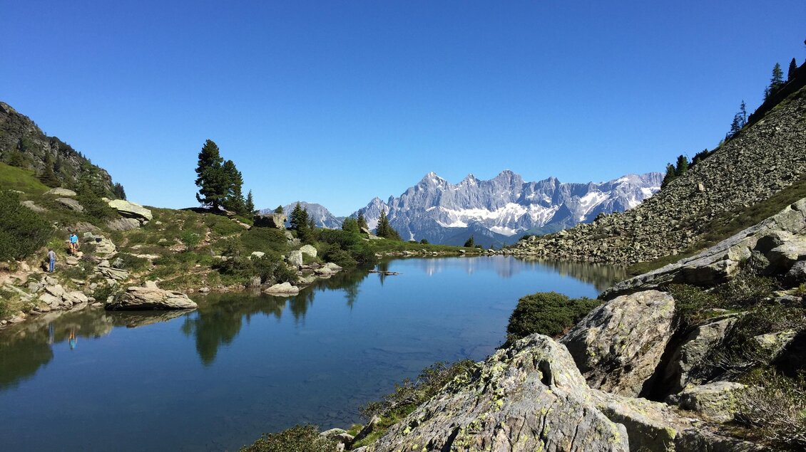 Eine ruhige Berglandschaft mit einem klaren See und hohen Gipfeln im Hintergrund. Der Himmel ist blau und die Natur ist üppig und grün. | © Markus Trinker