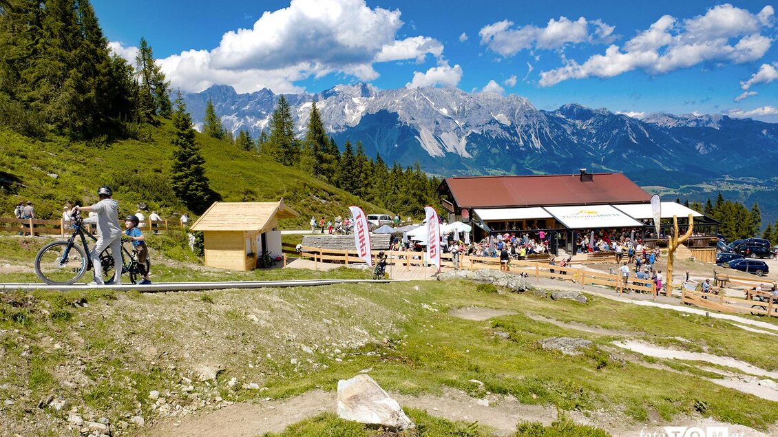 Eine malerische Berglandschaft mit einem Restaurant und vielen Besuchern. Radfahrer und Wanderer genießen die Natur unter einem blauen Himmel. | © Markus Trinker