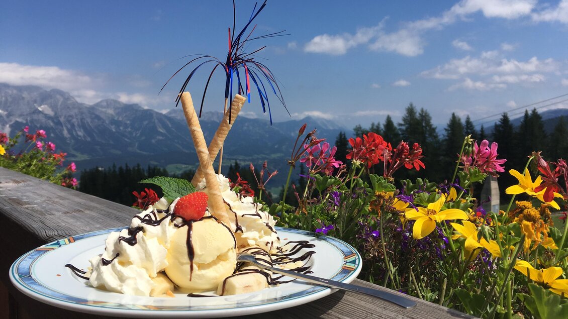 Ein leckeres Eisgericht mit Sahne und Schokoladensoße. Im Hintergrund blühen bunte Blumen vor einer beeindruckenden Bergkulisse. | © Reiteralmhütte 