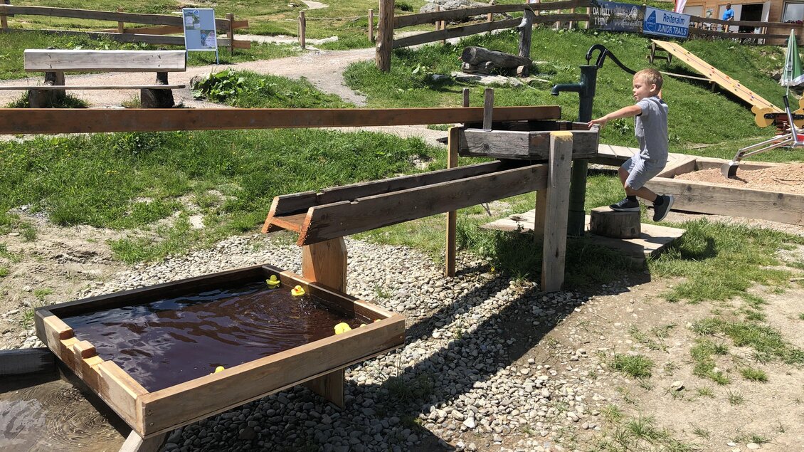 Ein Kind spielt an einer Wasserbahn mit Holzrutschen und Gummientchen. Im Hintergrund sieht man eine grüne Wiese und einige Gebäude. | © Markus Trinker