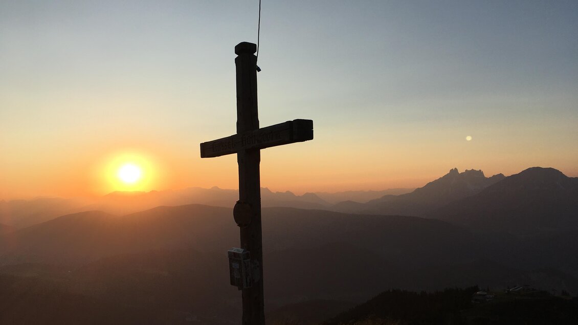 Ein Kreuz steht auf einem Berggipfel im Sonnenuntergang. Die Landschaft zeigt sanfte Hügel und Berge im Hintergrund. | © Reiteralmhütte 