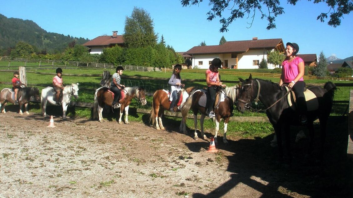Eine Gruppe von Kindern reitet auf Ponys und Pferden in einem Reitstall. Im Hintergrund sind grüne Wiesen und ein blauer Himmel zu sehen.