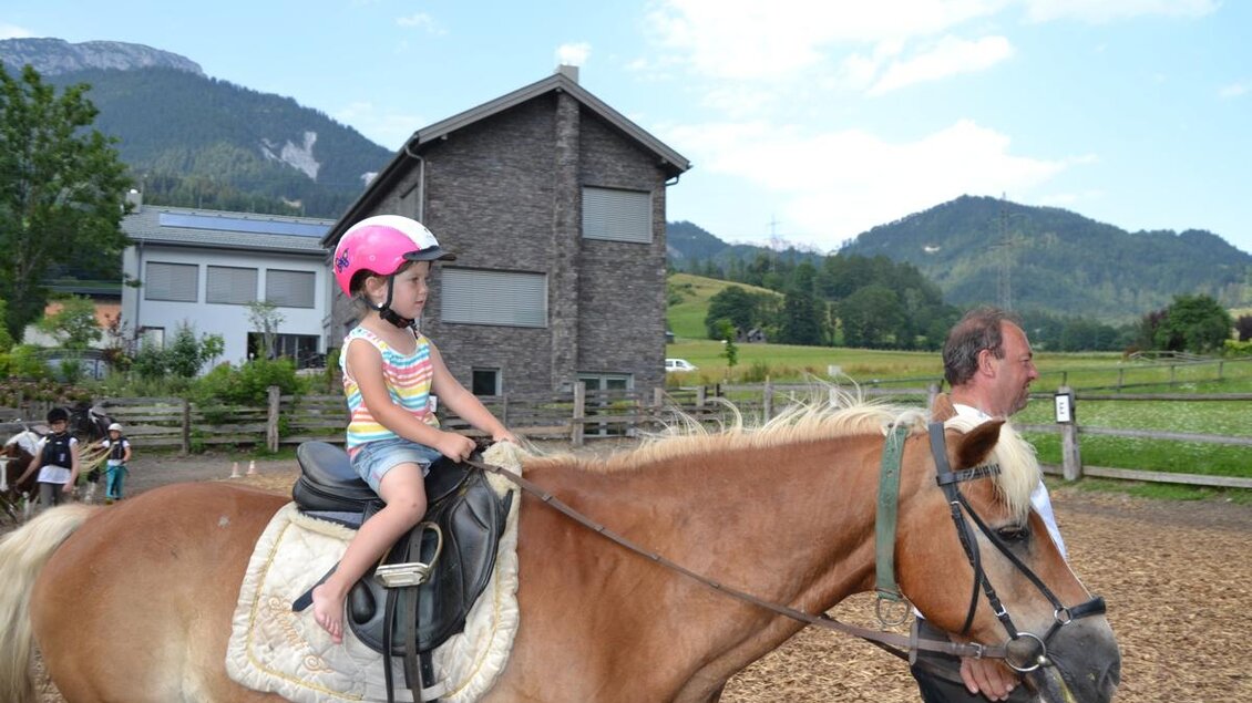Ein Mädchen reitet auf einem Pferd und trägt einen Helm. Im Hintergrund sieht man eine ländliche Umgebung mit Bergen und einem Haus.