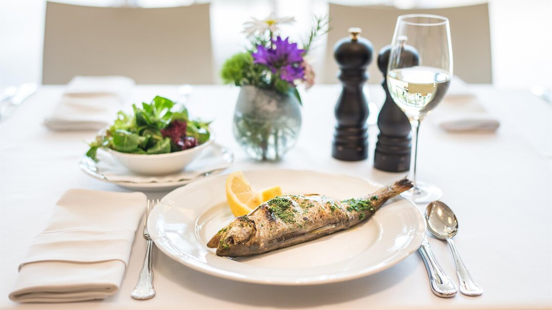 Ein eleganter Tisch mit einem gekochten Fisch, Zitrone und frischem Salat. Im Hintergrund steht ein Glas Weißwein und eine Blumenvase. | © Tauroa GmbH