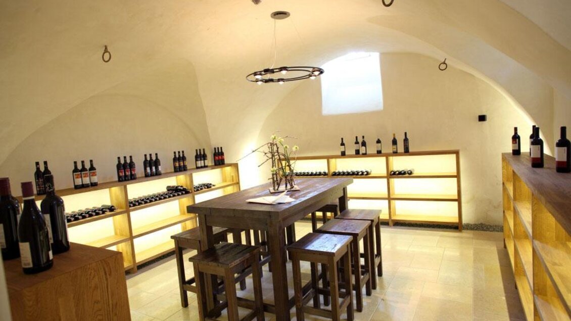 Ein Weinkeller mit Holzregalen, die mit Weinflaschen gefüllt sind. In der Mitte steht ein Holztisch mit Stühlen. | © Tauroa GmbH