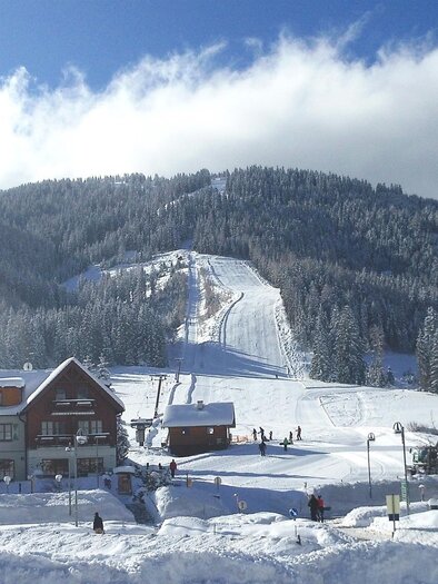 A snowy winter landscape with a ski resort and a cozy building in the foreground. In the background, there are snow-covered mountains and a clear blue sky. | © Passhöhe