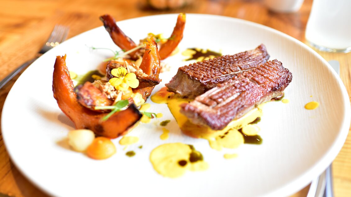 Ein kunstvoll angerichteter Teller mit gebratenem Fisch und Kürbisstücken. Das Gericht ist mit bunten Saucen und essbaren Blüten dekoriert. | © Erlebnisregion Murtal
