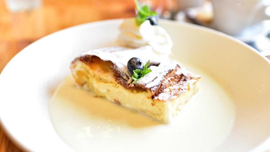 Ein Stück köstlicher Kuchen mit einer cremigen Füllung, garniert mit einer Sahnehaube und einer Blaubeere. Der Kuchen liegt in einer hellen Sauce und wird auf einem weißen Teller serviert. | © Erlebnisregion Murtal