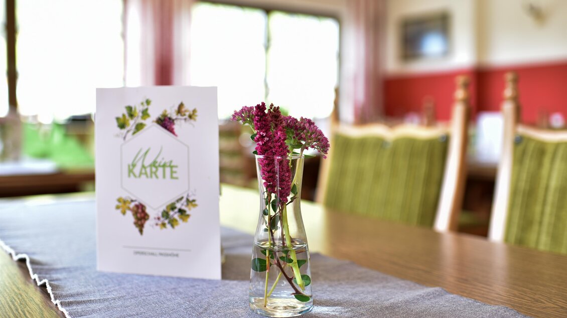 Ein Tisch in einem Restaurant mit einer Speisekarte und einer kleinen Vase voller Blumen. Der Hintergrund zeigt Stühle und Fenster mit natürlichem Licht. | © Erlebnisregion Murtal