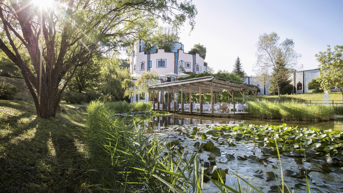 Ein ruhiger Teich mit Seerosen und üppigem Grün. Im Hintergrund befindet sich ein charmantes Gebäude und eine Terrasse mit Sitzgelegenheiten. | © Rogner Bad Blumau © Hundertwasser Architekturprojekt