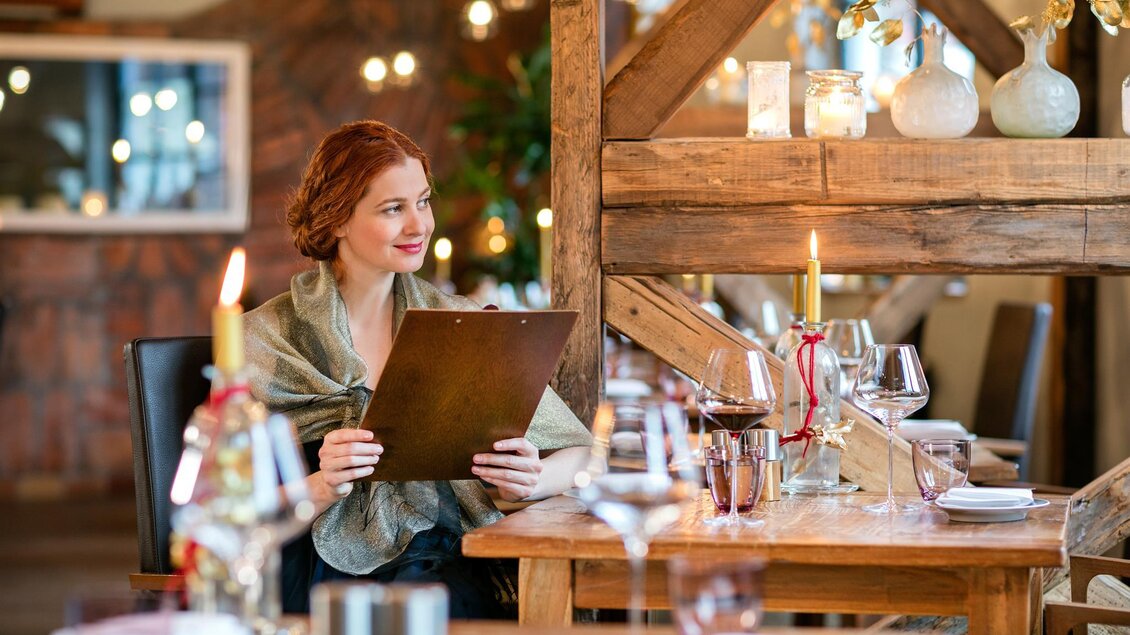 Eine Frau sitzt in einem elegant dekorierten Restaurant und schaut auf die Speisekarte. Der Tisch ist mit Gläsern und Kerzen liebevoll eingedeckt. | © RognerBadBlumau © Hundertwasser Architekturprojekt