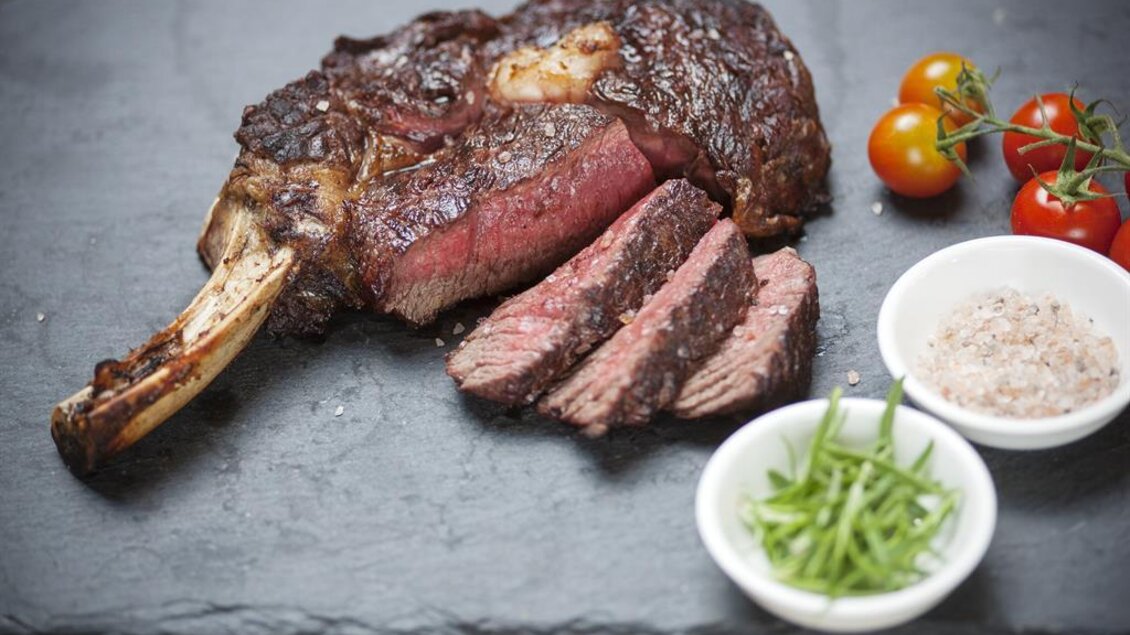 Ein köstliches Stück Fleisch auf einem schwarzen Steinbrett, perfekt zubereitet und in Scheiben geschnitten. Dazu frische Kirschtomaten und kleine Schalen mit Salz und Kräutern. | © Rogner Bad Blumau