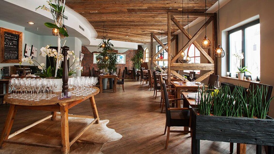 Ein modernes Restaurant mit Holzdekor und stilvollen Möbeln. Große Fenster lassen viel Licht hinein und tropische Pflanzen sorgen für eine einladende Atmosphäre. | © Rogner Bad Blumau