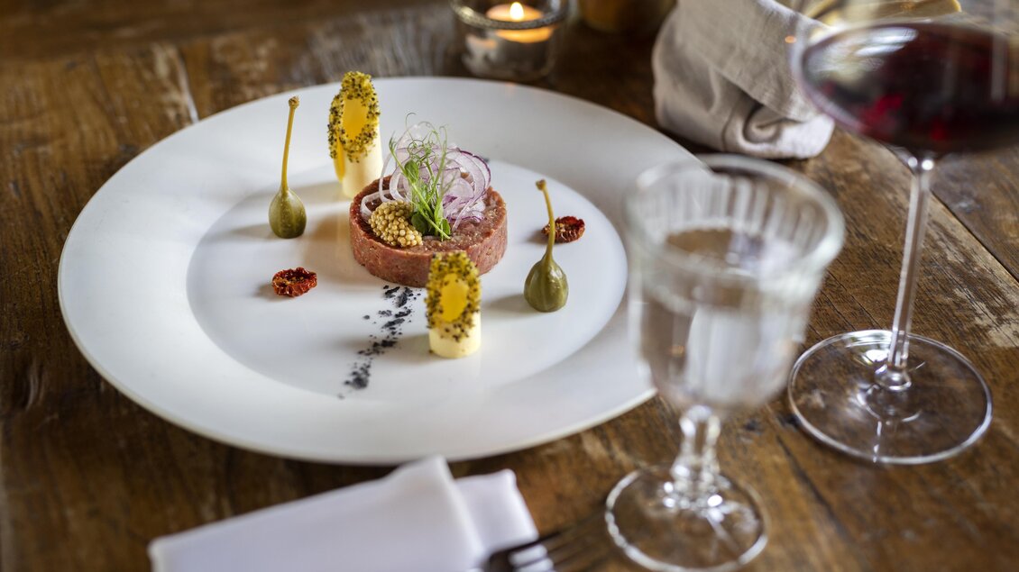 Ein sorgfältig angerichteter Teller mit einer Fleischpastete, garniert mit essbaren Blüten und grünen Pfefferkörnern. Daneben ein Glas Wasser und ein Rotwein in einem eleganten Restaurant. | © Rogner Bad Blumau © Hundertwasser Architekturprojekt