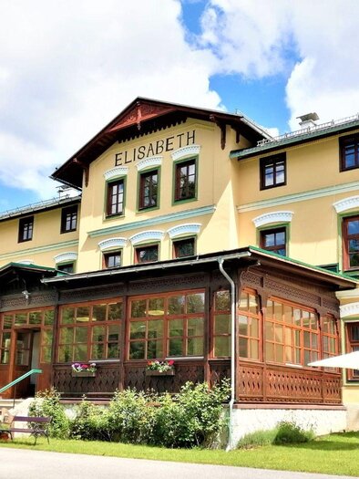 Ein historisches Hotel mit gelber Fassade und einer schönen Veranda. Umgeben von grüner Landschaft und mit Tischen im Freien. | © Gasthof Elisabeth