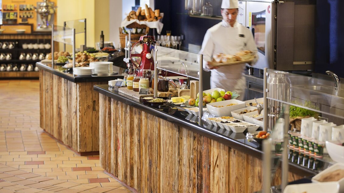 Ein Buffet mit einer Vielzahl von Speisen und Getränken ist zu sehen. Ein Koch bereitet gerade die Gerichte vor. | © Rogner Bad Blumau