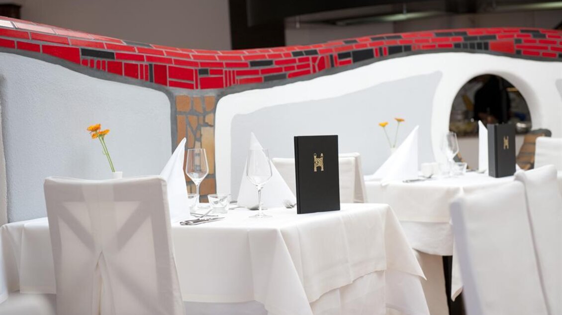Ein elegantes Restaurant mit weißen Tischen und Stühlen. Die Tische sind mit weißen Tischdecken und Menükarten dekoriert, und eine Blume bringt Farbe ins Ambiente. | © Rogner Bad Blumau