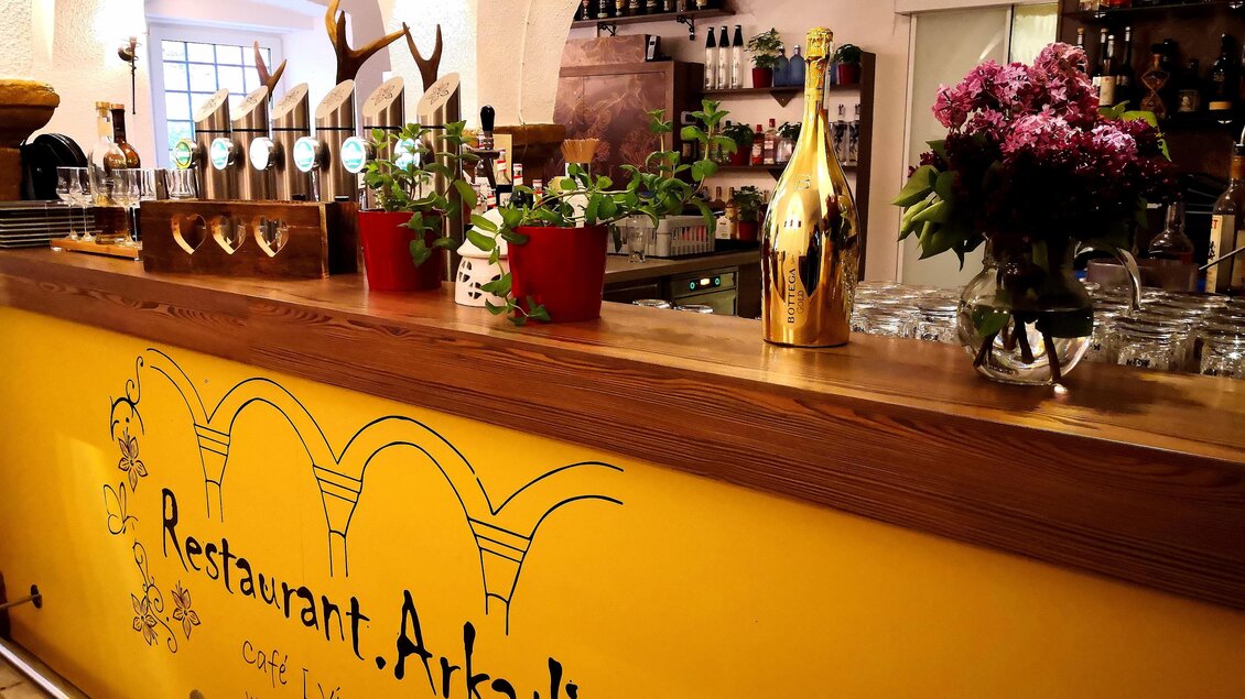 Eine einladende Bar im Restaurant Arkadia mit einer gelben Wand und Pflanzen. Auf der Theke stehen Gläser und eine goldene Flasche. | © Arkadia
