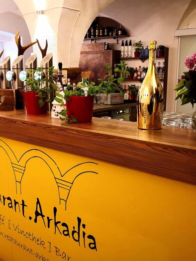 An inviting bar in the Arkadia restaurant with a yellow wall and plants. Glasses and a golden bottle are on the counter. | © Arkadia