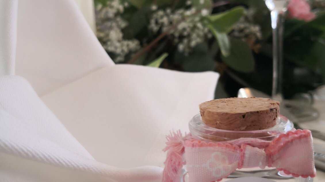 Ein dekoratives Glas mit einem Korken und einem süßen Aufkleber steht auf einem Tisch. Im Hintergrund sind Blumen und eine Weinglashalterung zu sehen. | © Arkadia