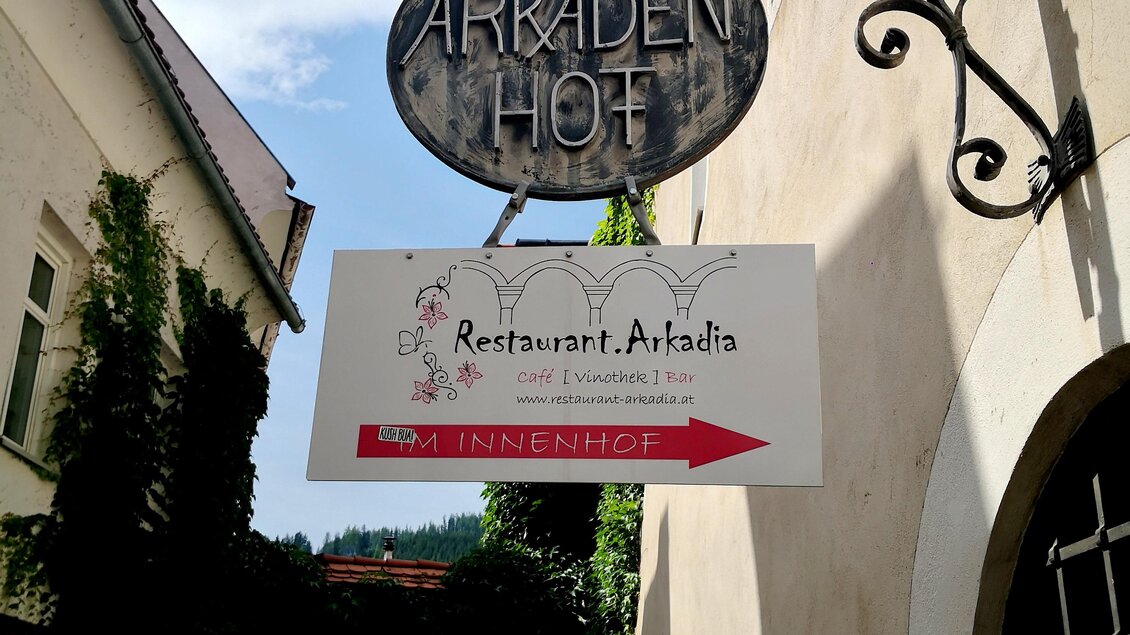 Ein Schild mit der Aufschrift „Arkaden Hof“ und „Restaurant Arkadia“ hängt an einem Gebäude. Im Hintergrund ist ein malerischer Weg mit Pflanzen zu sehen. | © Arkadia