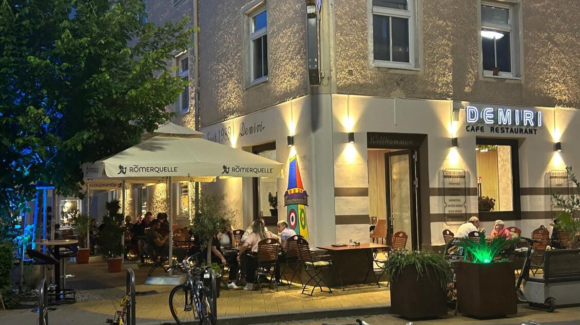 Außenansicht des Demiri Café-Restaurants bei Nacht mit beleuchtetem Eingangsbereich und Gästen. | © Demiri